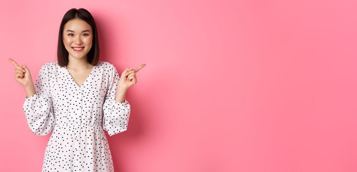 Beautiful asian woman making choice on shopping, pointing fingers sideways and showing variants, smiling at camera, standing over pink background
