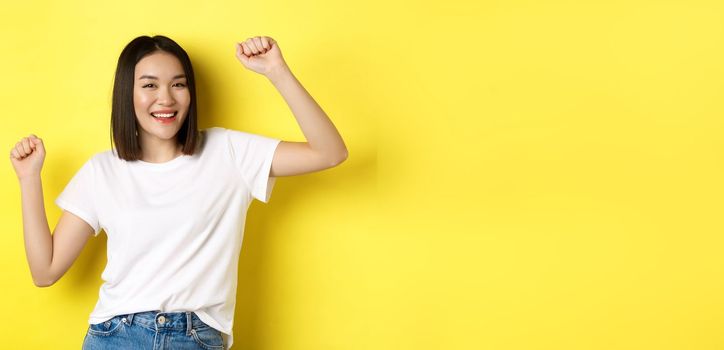 Carefree asian girl dancing and having fun, posing in white t-shirt against yellow background