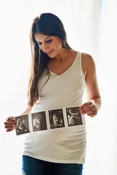 Hes got his mothers good looks already. a happy pregnant woman holding up a series of ultrasound pictures in front of a window at home.
