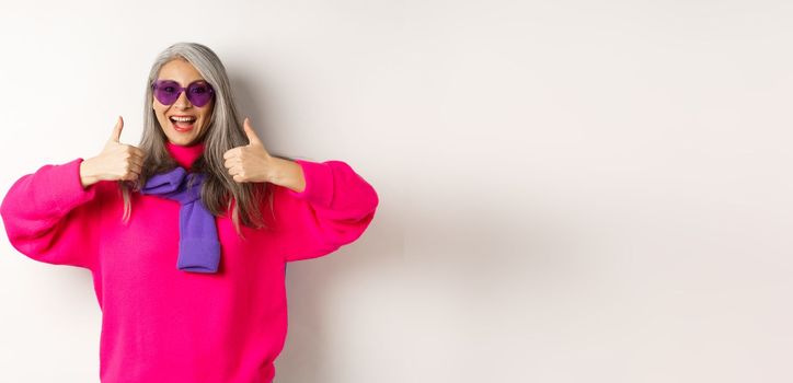 Shopping concept. Stylish asian senior woman in sunglasses and trendy outfit, showing thumb-up in approval, recommending shop, standing over white background