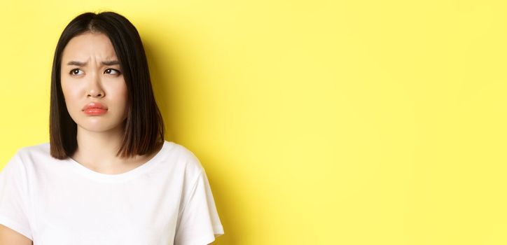 Sad and jealous asian girl sulking, frowning and looking left with upset face, standing over yellow background