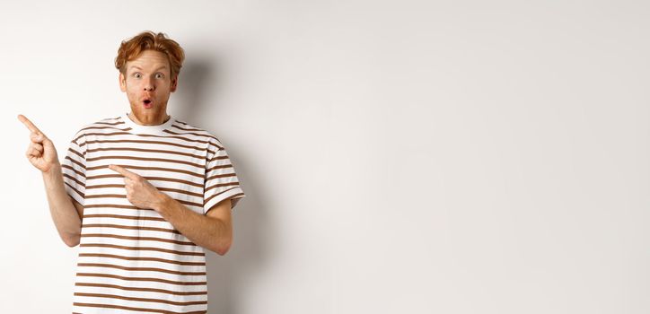 Amazed man with ginger hair checking out promo offfer, pointing left at logo and smiling at camera, standing over white background