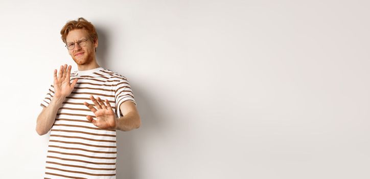 Displeased young man with red hair and bristle showing stop gesture, refusing something bad with disgusted face, standing over white background