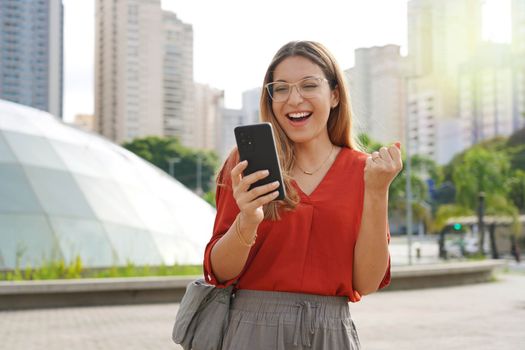 Brazilian surprised girl watching joyful her smartphone with modern sustainable city on the background