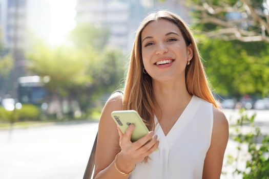 Brazilian confident businesswoman looking at camera using smartphone outdoors on summer. Copy space.