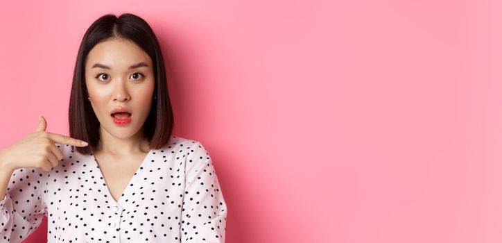 Image of confused asian woman pointing at herself, drop jaw and staring questioned at camera, standing over pink background