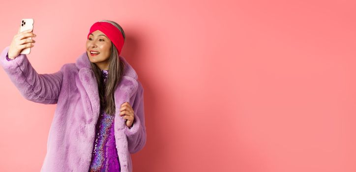 Fashion concept. Beautiful asian senior lady taking selfie on smartphone, posing in party glittering dress and purple faux coat, standing over pink background