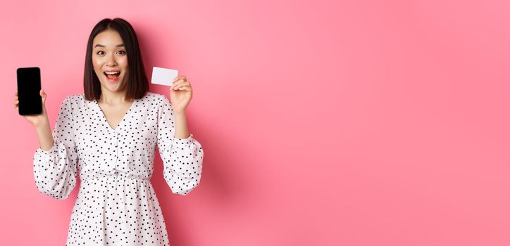 Cute asian woman shopping online, showing bank credit card and mobile screen, smiling and looking at camera, standing over pink background
