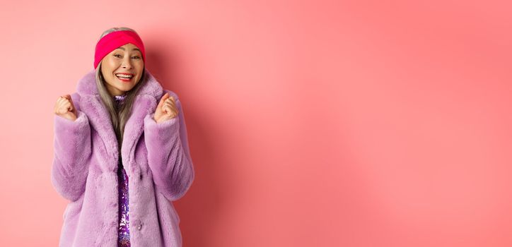Cheerful asian lady in stylish funky coat, celebrating victory or success, saying yes and smiling happy, standing over pink background