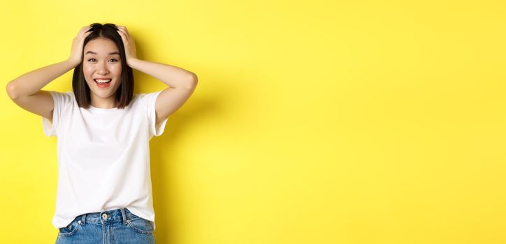 Amazed asian girl winning prize and looking with excitement and disbelief, holding hands on head and smiling happy, standing over yellow background