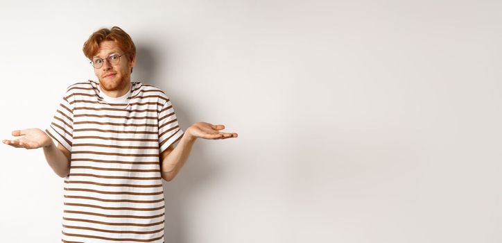 Confused young man with red hair, shrugging shoulders and looking indecisive, standing over white background