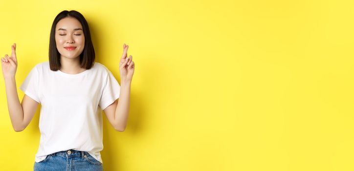 Hopeful asian girl making wish, cross fingers for good luck and praying with eyes closed, saying please, standing over yellow background