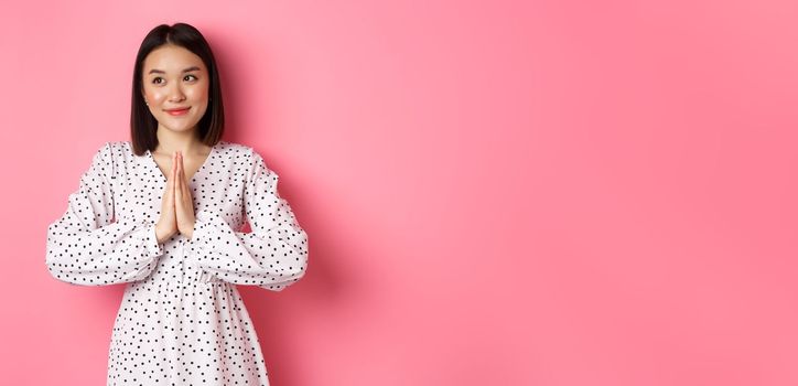 Beautiful angelic asian woman smiling, holding hands in pray and looking left at copy space with innocent cute gaze, standing over pink background
