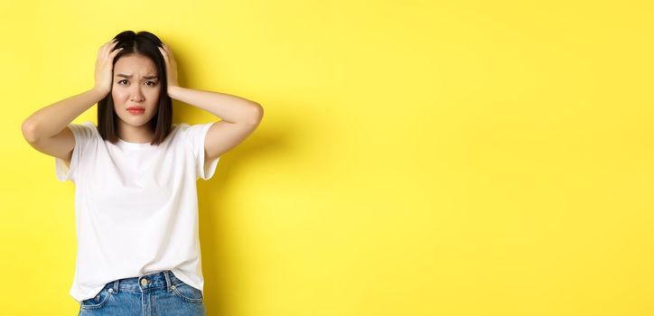 Asian woman holding hands on head and looking sad, having a problem, standing anxious against yellow background