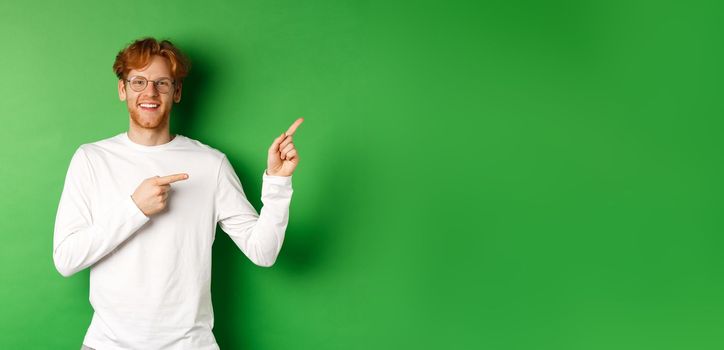 Handsome young man with ginger hair and glasses, pointing fingers right at copy space and smiling, standing over green background