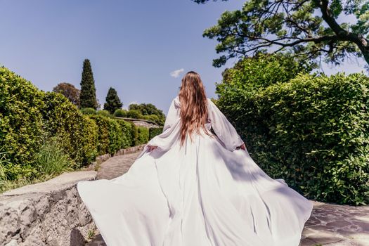 Brunette runs white dress park. A beautiful woman with long brown hair and a long white dress runs along the path along the beautiful bushes in the park, rear view
