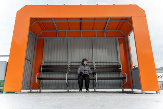 a lonely child waiting for a bus at an empty bus stop in winter. A small orange bus stop and a waiting boy sitting on a bench on a cold winter day.