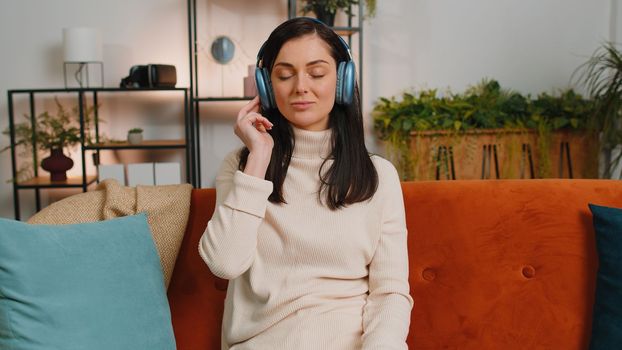 Happy young woman in wireless headphones relaxing sitting on sofa at home listening favorite music