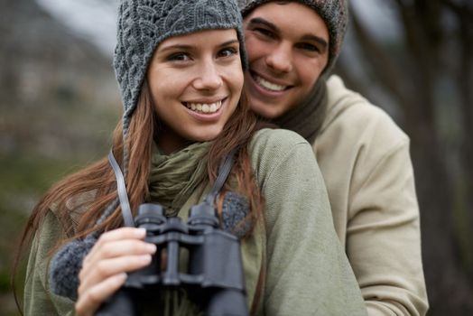 What a great view. A young couple smiling at the camera as they hold a pair of binoculars on a mountainside.