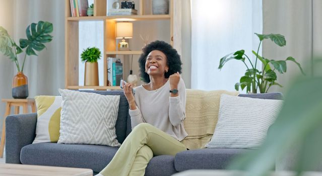 African woman celebrating a new job while sitting at home on a couch. A young females loan is approved via an email on her phone. A happy and excited lady cheering for a promotion on a sofa