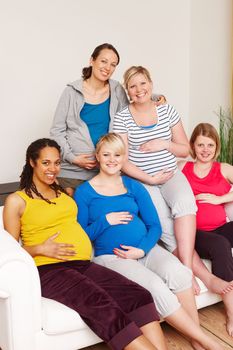 We cant wait for the big day. A group of multi-ethnic pregnant women sitting on a couch and smiling at the camera.