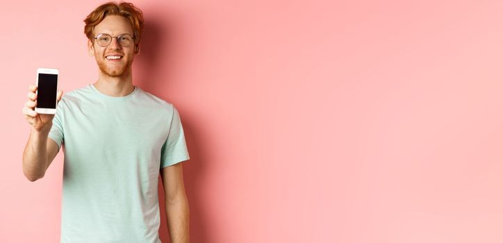 Handsome redhead man in glasses showing blank smartphone screen and smiling, demonstrate online promo or application, standing over pink background