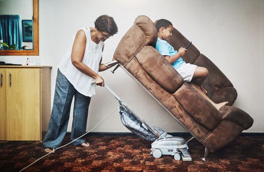 Grandmas got to get her cleaning done. a grandmother vacuuming under a couch which her grandson is lying on at home.