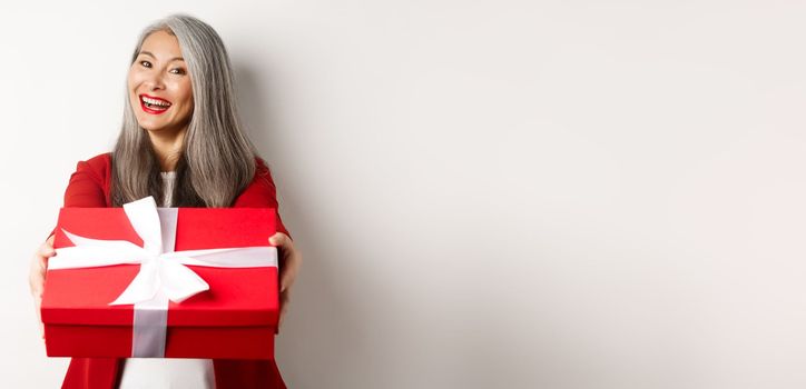 Happy holidays concept. Cheerful asian senior woman giving you red gift box and smiling, congratulating with valentine day, standing over white background