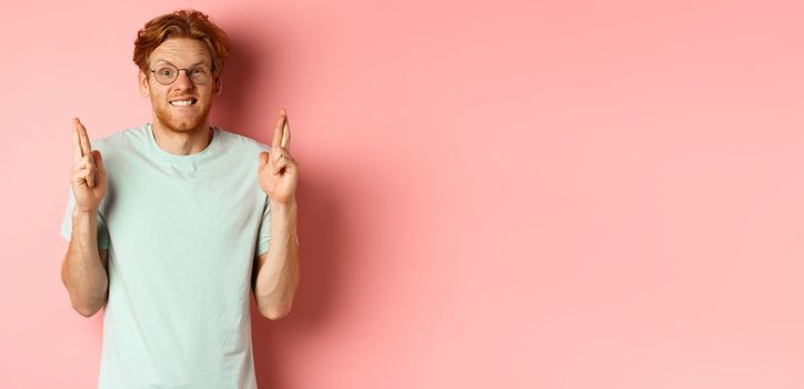 Worried redhead man waiting for results, expecting something with fingers crossed, biting finger and looking at something risky, standing over pink background