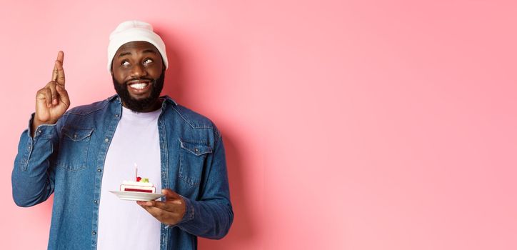 Hopeful Black guy celebrating birthday, making wish with fingers crossed, holding bday cake with candle, standing against pink background