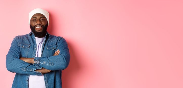 Handsome hip hop style Black man in beanie and denim shirt, smiling confident, cross arms on chest and staring at camera on pink background