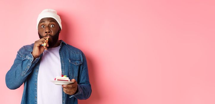 Happy Black hipster celebrating birthday, blowing party whistle, holding bday cake with candle, standing over pink background