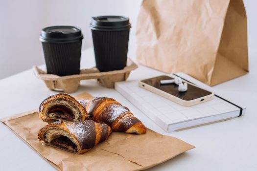 Chocolate croissants with cups of coffee and notepad with smartphone and earphones on the white table, food delivery concept