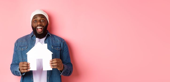 Real estate concept. Happy smiling african-american man holding house model, looking excited at camera, searching for home, standing over pink background