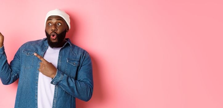 Impressed hipster black man in beanie and denim shirt, looking and pointing at upper left corner logo, staring amazed, standing over pink background