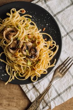 Spaghetti pasta with champignon mushrooms sprinkled with cheese parmesan and green onions on a black plate with a towel and a fork on a wooden table