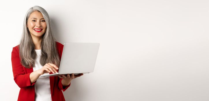 Business. Senior woman working on laptop, wearing office outfit and smiling, standing over white background