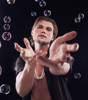 Man, studio portrait and bubbles with for magic, art performance or creative show by black background. Model, hands and soap with rainbow light, dark mystery aesthetic and sexy fashion by backdrop