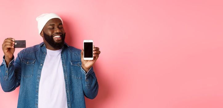 Online shopping. Satisfied Black man nod in approval, smiling and looking at phone, showing credit card and smartphone screen, standing over pink background