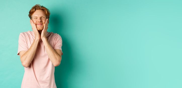 Image of cute and silly young man with red hair, feeling satisfaction while touching his face, smiling happy, standing over turquoise background