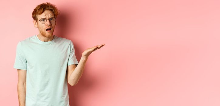 Portrait of handsome redhead man in glasses looking confused, open mouth and stare at camera, cant understand nothing, standing over pink background