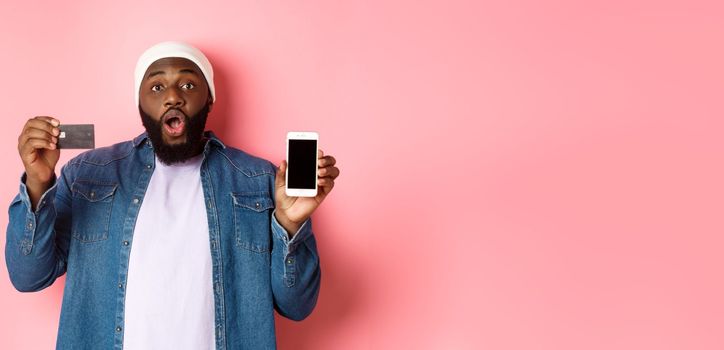 Online shopping. Excited Black man showing credit card and mobile phone screen, standing over pink background amazed