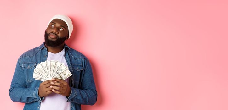 Dreamy african-american man thinking about shopping, holding money dollars and looking upper left corner, standing over pink background
