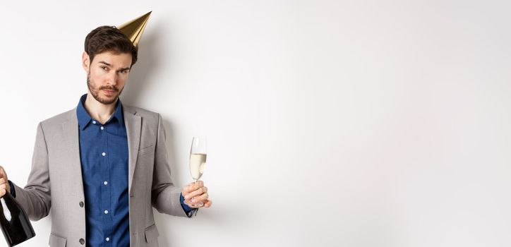 Party guy standing in birthday hat and celebrating, holding champagne bottle and glass, looking drunk, wearing grey suit