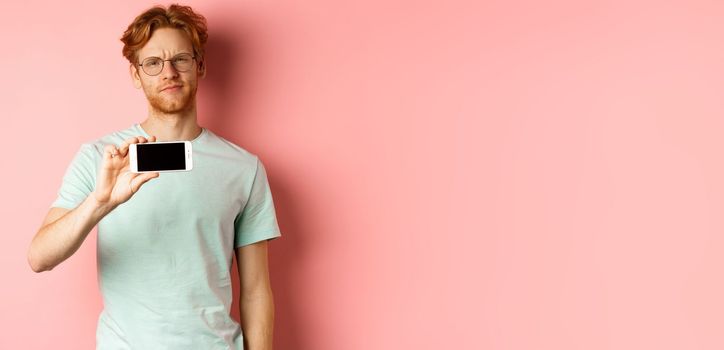 Skeptical redhead guy in glasses showing smartphone screen horizontally, smirk and frowning disappointed, standing over pink background
