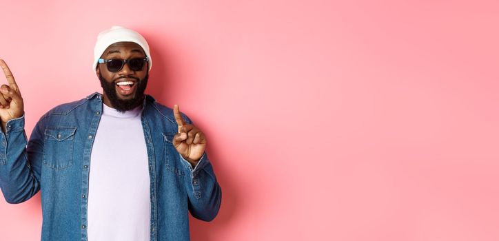 Happy bearded african-american man showing announcement, pointing fingers upper left corner and smiling, standing in beanie and sunglasses over pink background