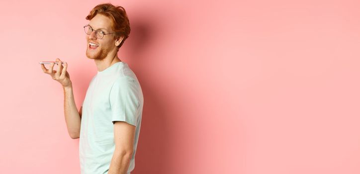 Handsome young man standing in profile and talking on speakerpone, record voice message, turn head at camera and smiling pleased, standing over pink background