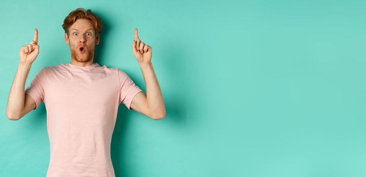 Impressed handsome guy with red hair pointing fingers up, demonstrate promo offer, standing in t-shirt over mint background