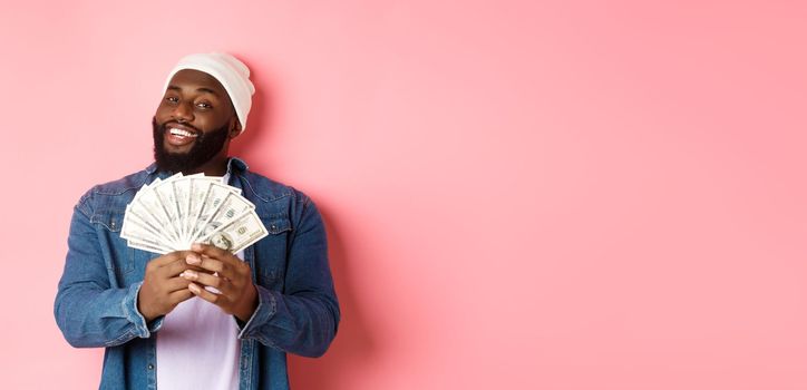 Satisfied rich african-american man showing money, smiling pleased and show-off with income, standing with dollars over pink background
