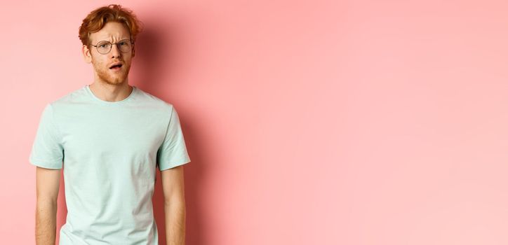 Portrait of handsome redhead man in glasses looking confused, open mouth and stare at camera, cant understand nothing, standing over pink background